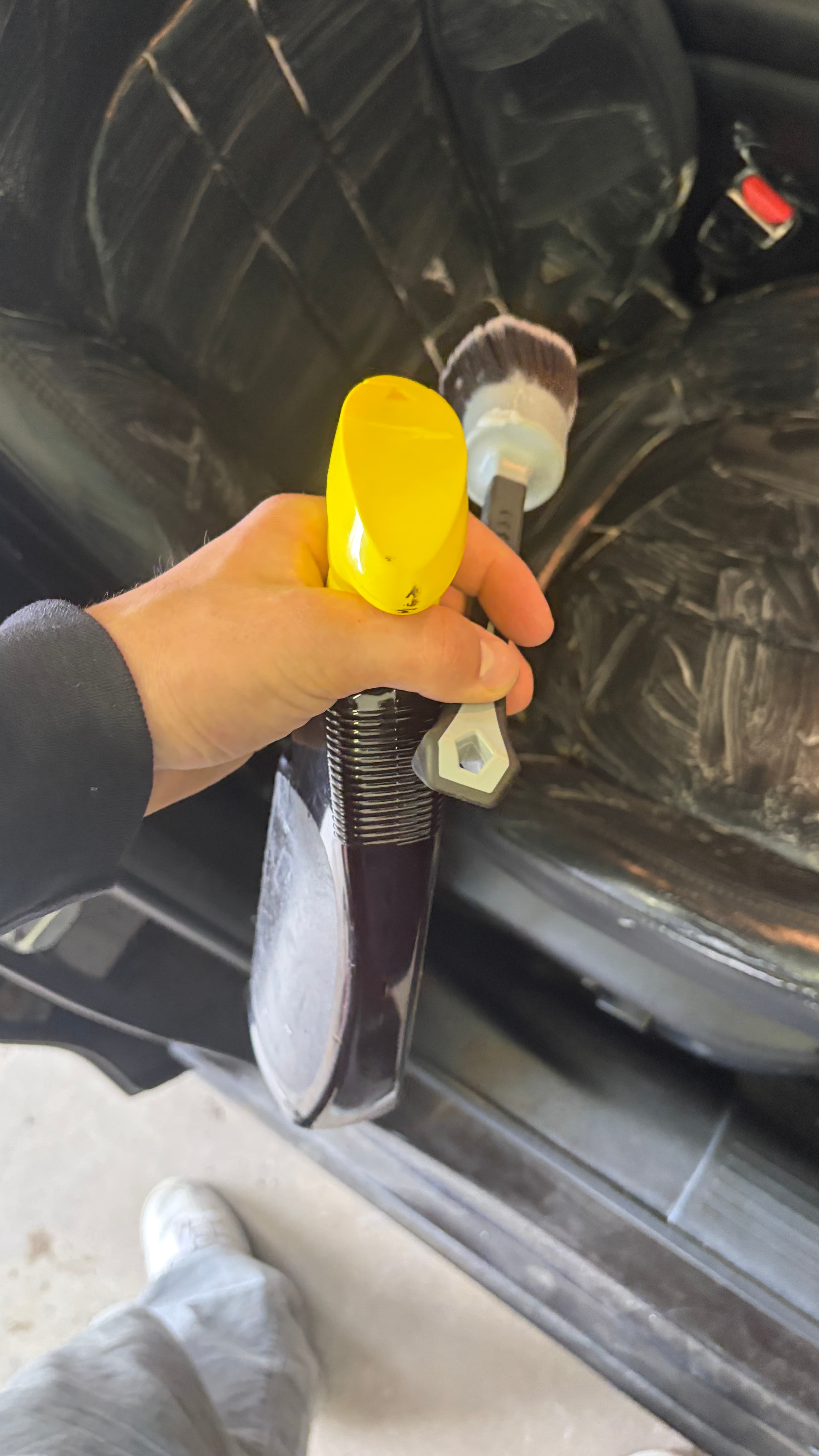 Hand holding a spray bottle and detailing brush over a leather car seat being cleaned.