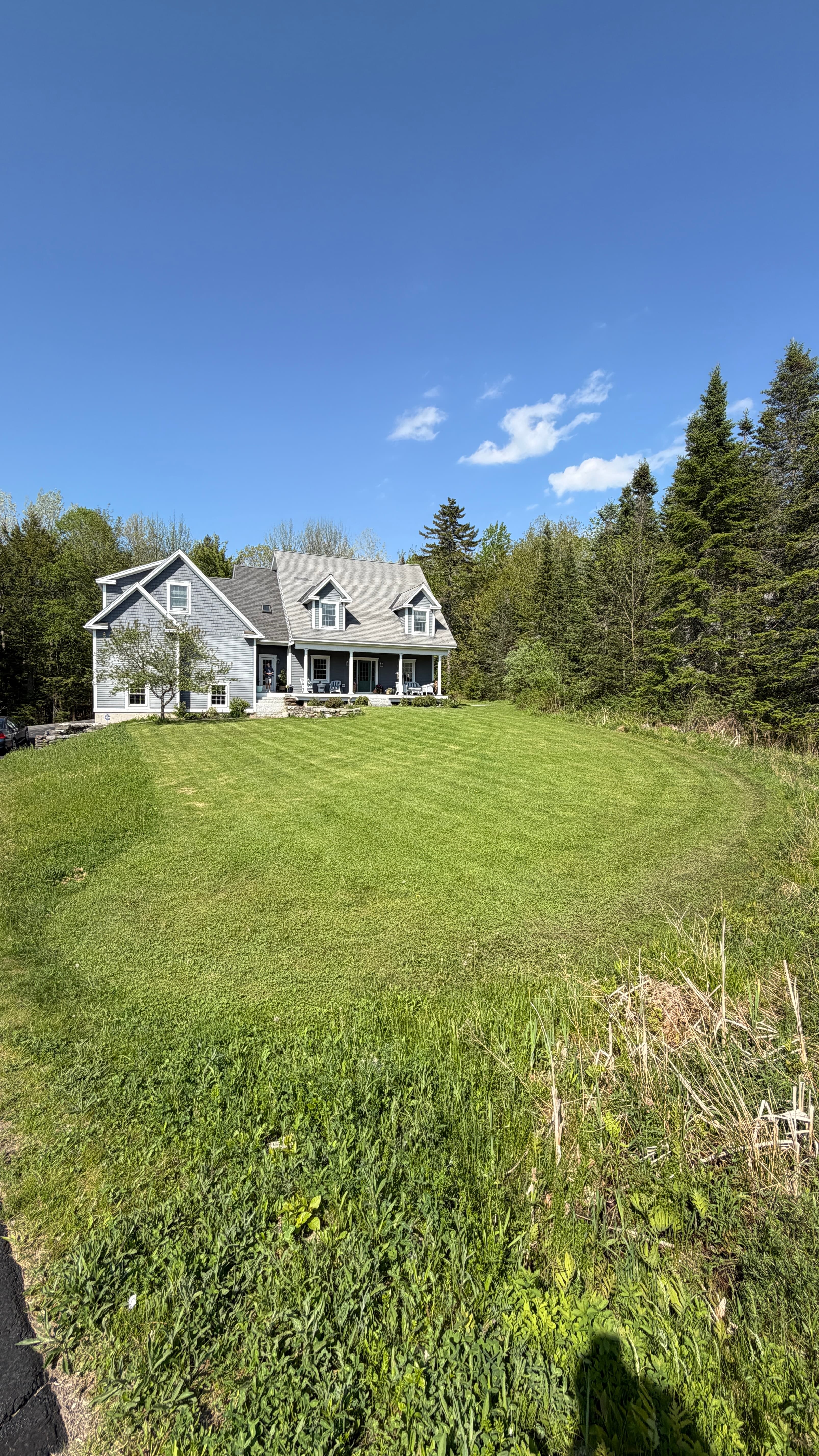 Grey house with white trim on a large green lawn surrounded by dense forest.