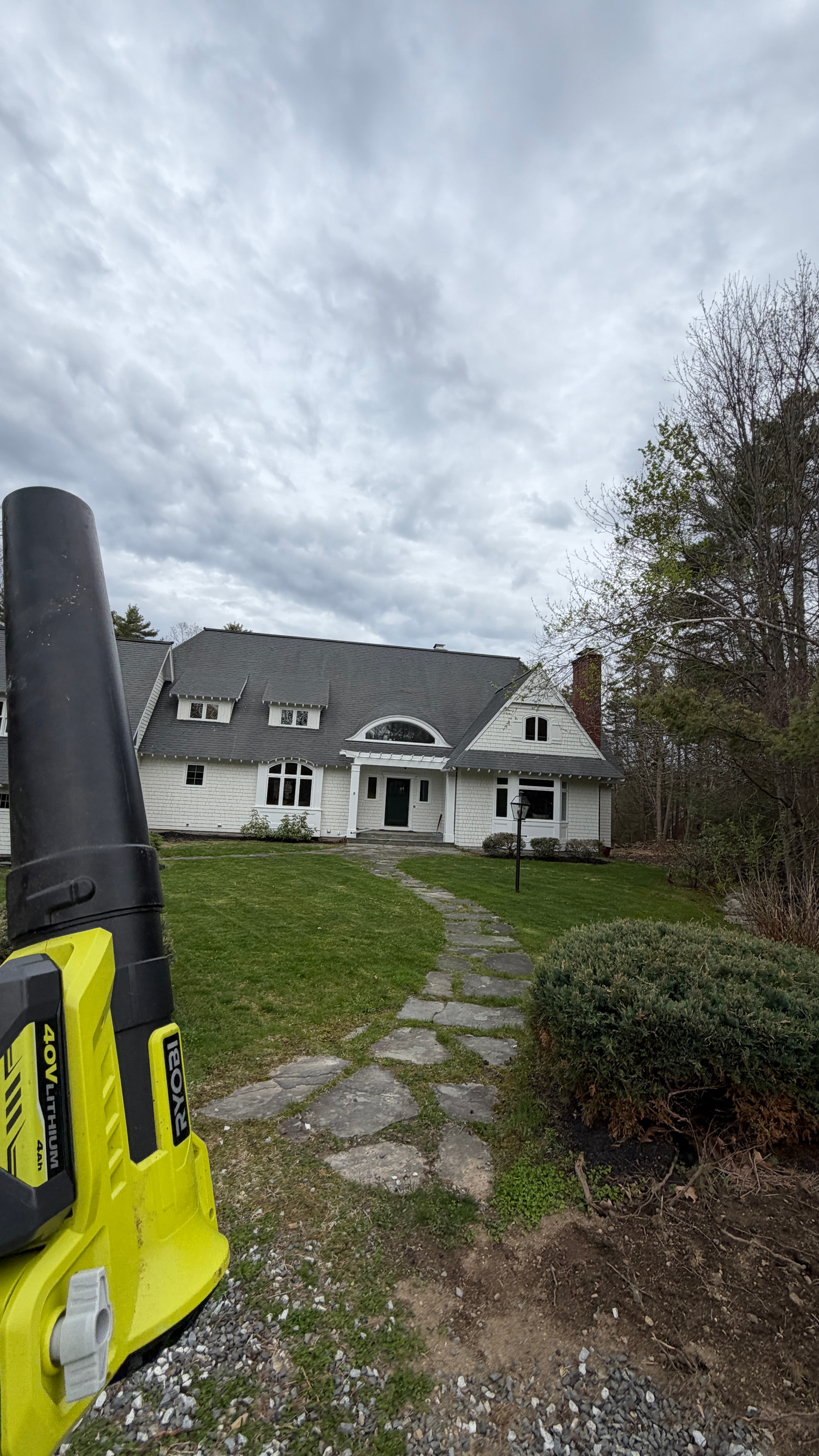 Ryobi leaf blower foreground, stone path leading to a white house under a cloudy sky.
