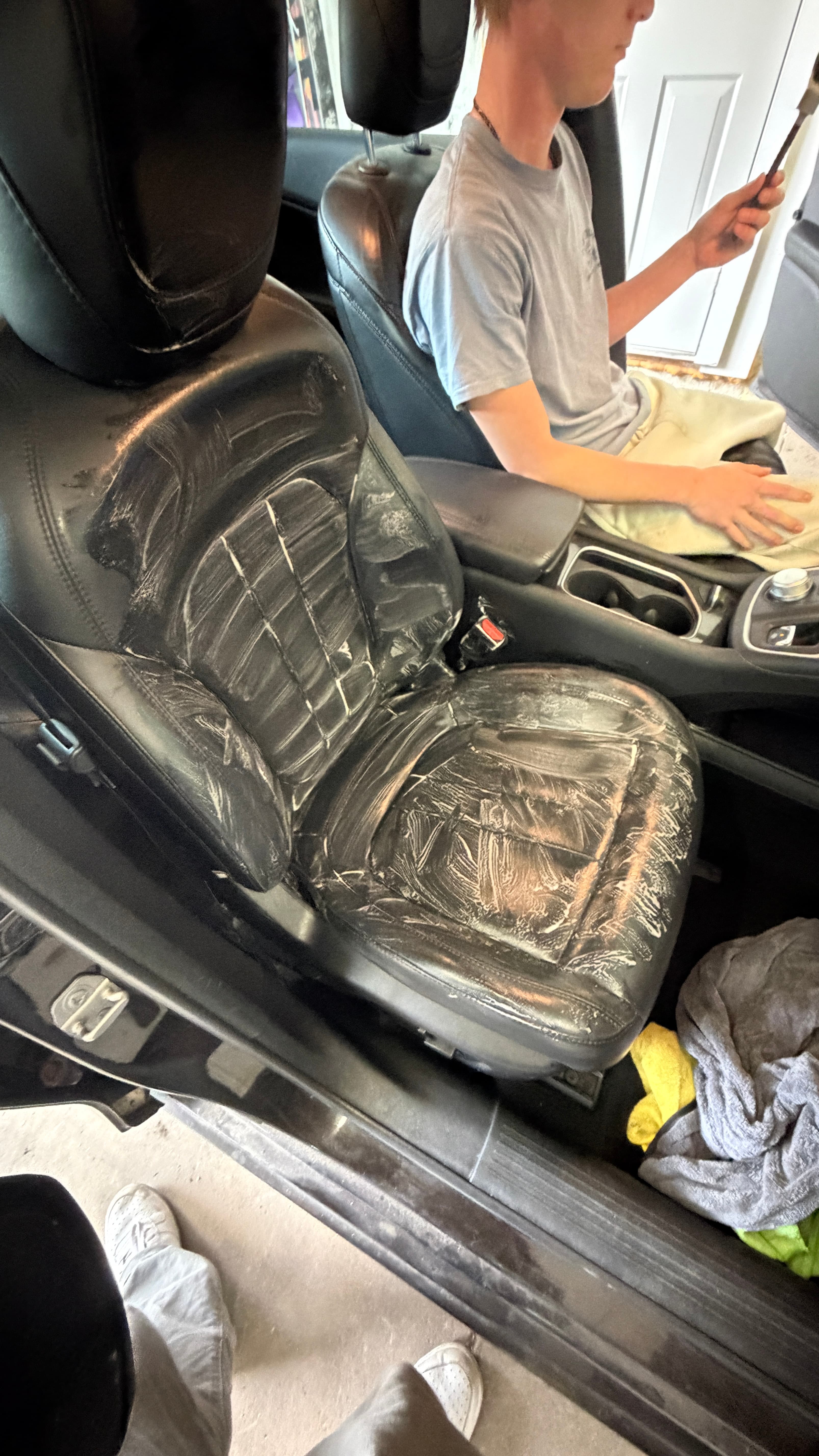 Black leather car seat covered in white cleaning cream with a person sitting nearby.