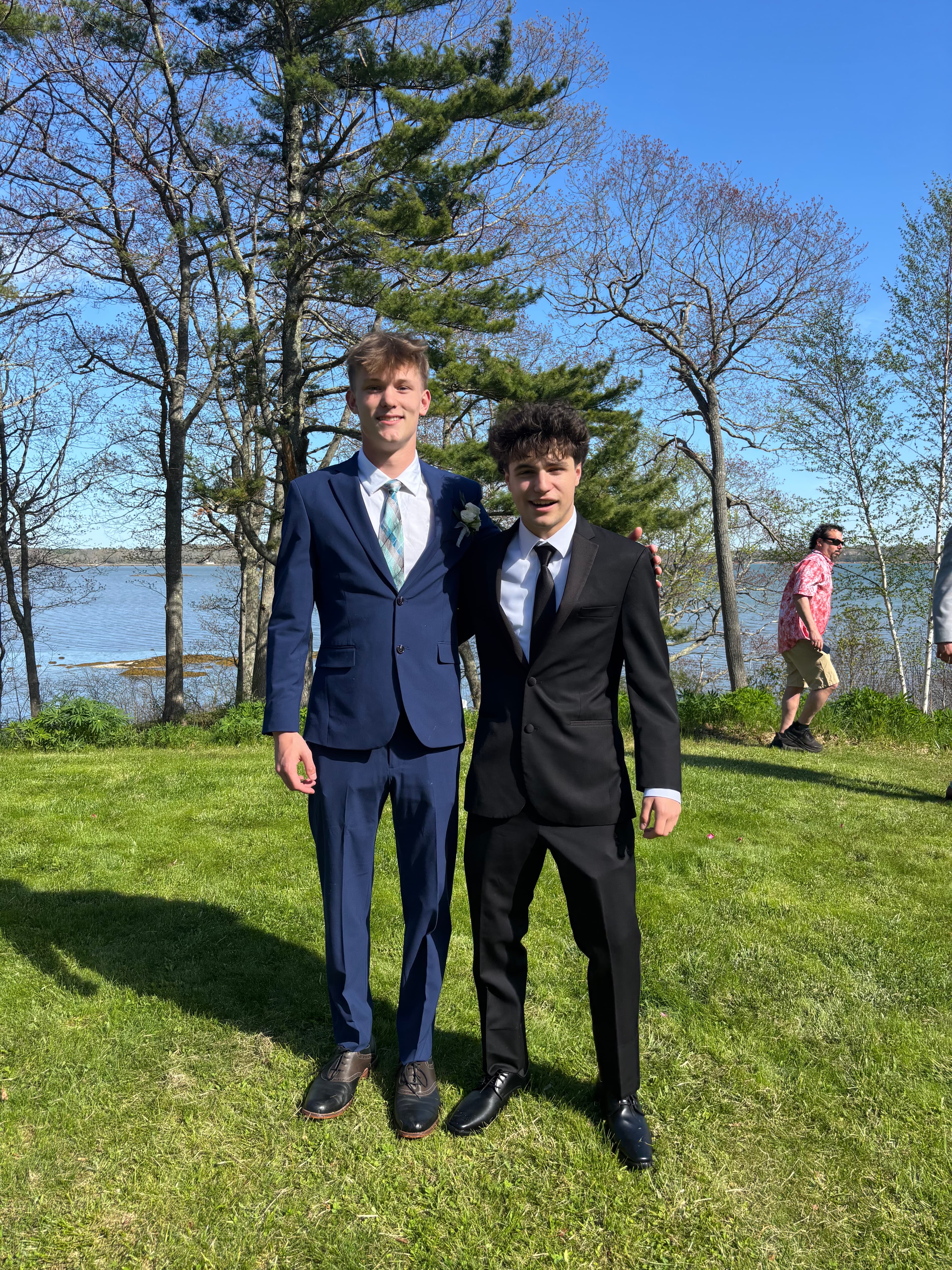 Two young men in blue and black suits pose on a lawn near a lake.
